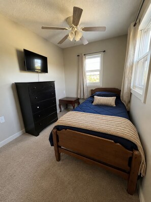 A bedroom with a bed, a dresser, a television, a ceiling fan, and a window with curtains.