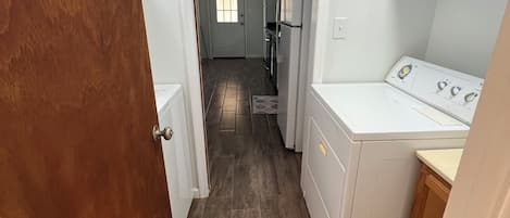 A narrow hallway with a wooden door on the left, a white laundry area on the right, and a view into a kitchen area at the end.