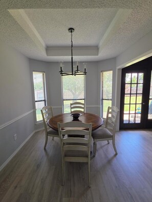 A dining room with a round table, four chairs, a chandelier, and large windows with blinds.