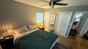 Een slaapkamer met een bed, een bedlampje, een plafondventilator en een deur naar een andere kamer.