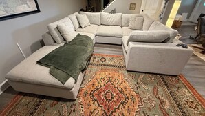 Een woonkamer met een hoekbank, een klein poufje, een kamerkleed en een trap naar een hoger gelegen verdieping.