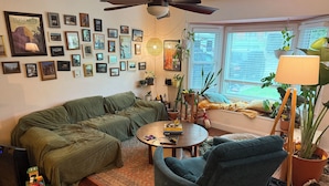Een woonkamer met een groene bank, een blauwe fauteuil, een klein tafeltje met boeken, een plafondventilator en een raam met zonwering.