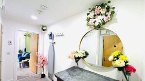 Ein Zimmer mit Holztür, einem Spiegel mit goldener Rahmung, einem Schminktisch und einer Vase mit Blumen.