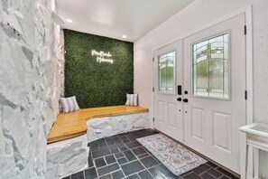 Ein modernes Interieur mit einer Steinwand, einer grünen Wand mit leuchtendem Neonzeichen, einer Holzbank, einer weißen Tür mit Glasfeldern und einem gemusterten Teppich.
