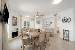 A modern living room with a fireplace, a dining table with chairs, a ceiling fan, and a wall clock.