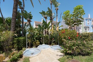 A garden with three circular water features, palm trees, and a building with a tiled roof in the background.