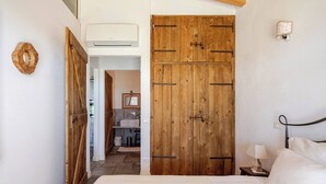 Ein Schlafzimmer mit Holzschrank, Bett, Nachttisch und einer Wandleuchte. Durch eine offene Tür ist ein Badezimmer zu sehen.