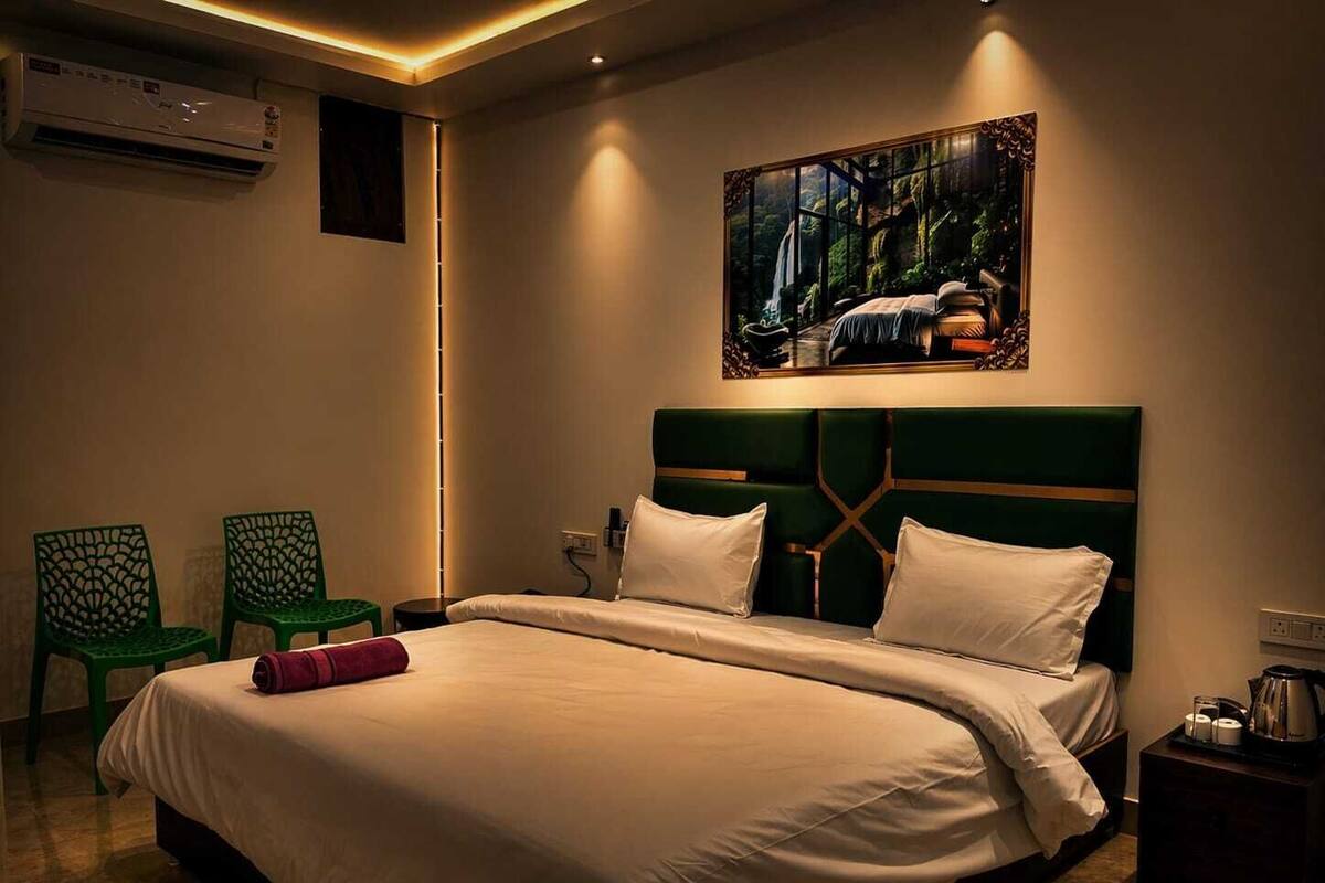 A hotel room with a large bed, a headboard, a nightstand, a green chair, a small table, and a painting on the wall.