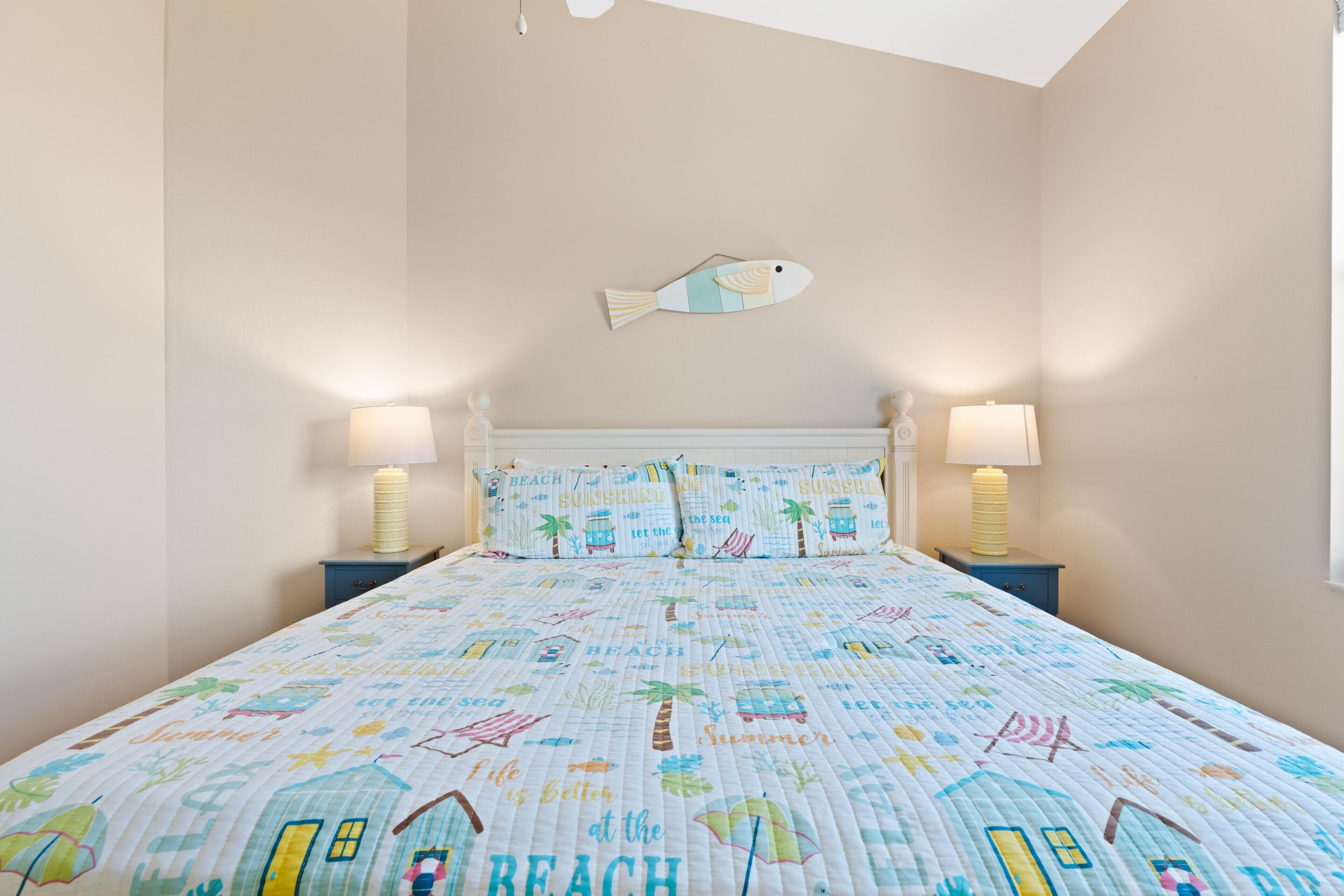 A bedroom with a beach-themed bedspread, two bedside lamps, a wall-mounted fish decoration, and a ceiling light fixture.