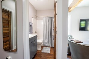 Une salle de bain avec une porte en bois, des toilettes blanches, un rideau de douche, un meuble-lavabo avec un miroir et un téléviseur à écran plat fixé au mur.