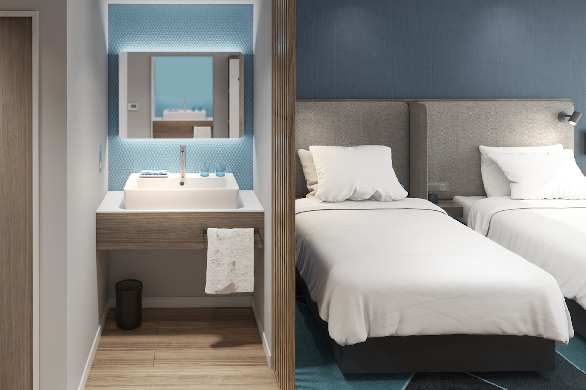 A hotel room with a wooden door, a bathroom with a sink and mirror, and two beds with white bedding.