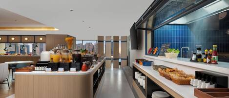 Un área de desayuno moderno en un hotel, con una isla central, barra libre y vistas a la ciudad.