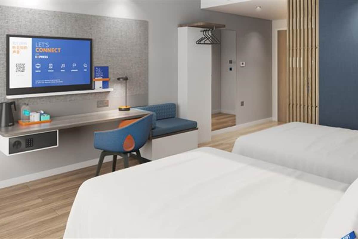 A modern hotel room with a bed, a desk with a computer, a chair, and a TV displaying a digital interface.