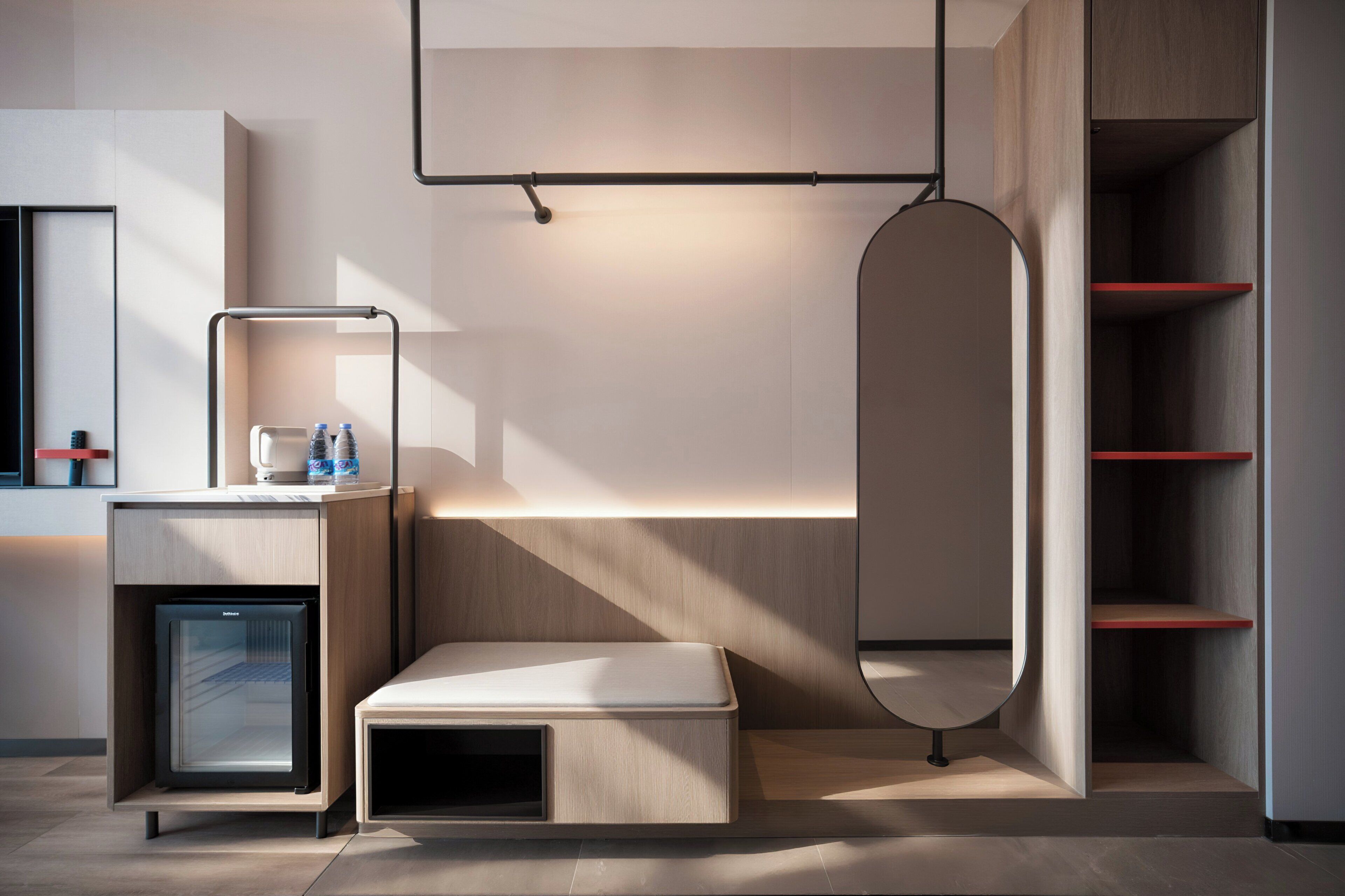 Moderni hotellihuone, jossa on sisäänrakennettu minijääkaappi, yöpöytä lampun kanssa, penkki ja suuri peili.