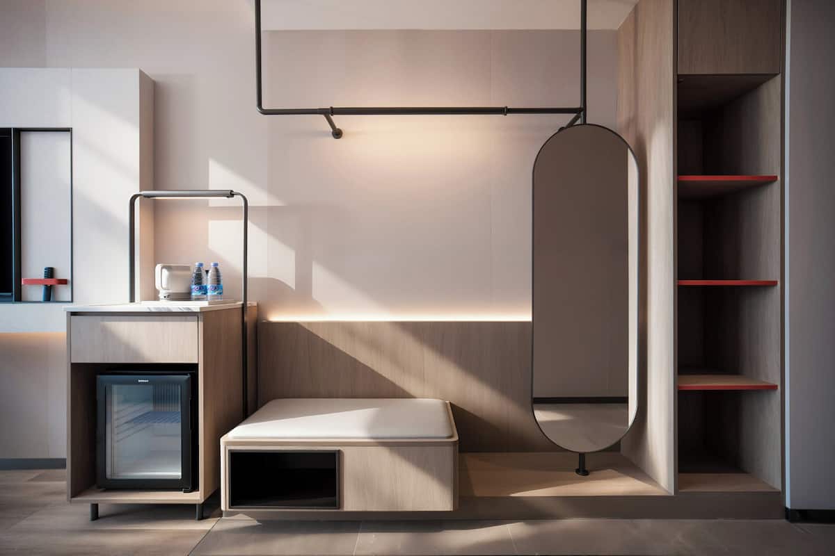 Moderni hotellihuone, jossa on sisäänrakennettu minijääkaappi, yöpöytä lampun kanssa, penkki ja suuri peili.