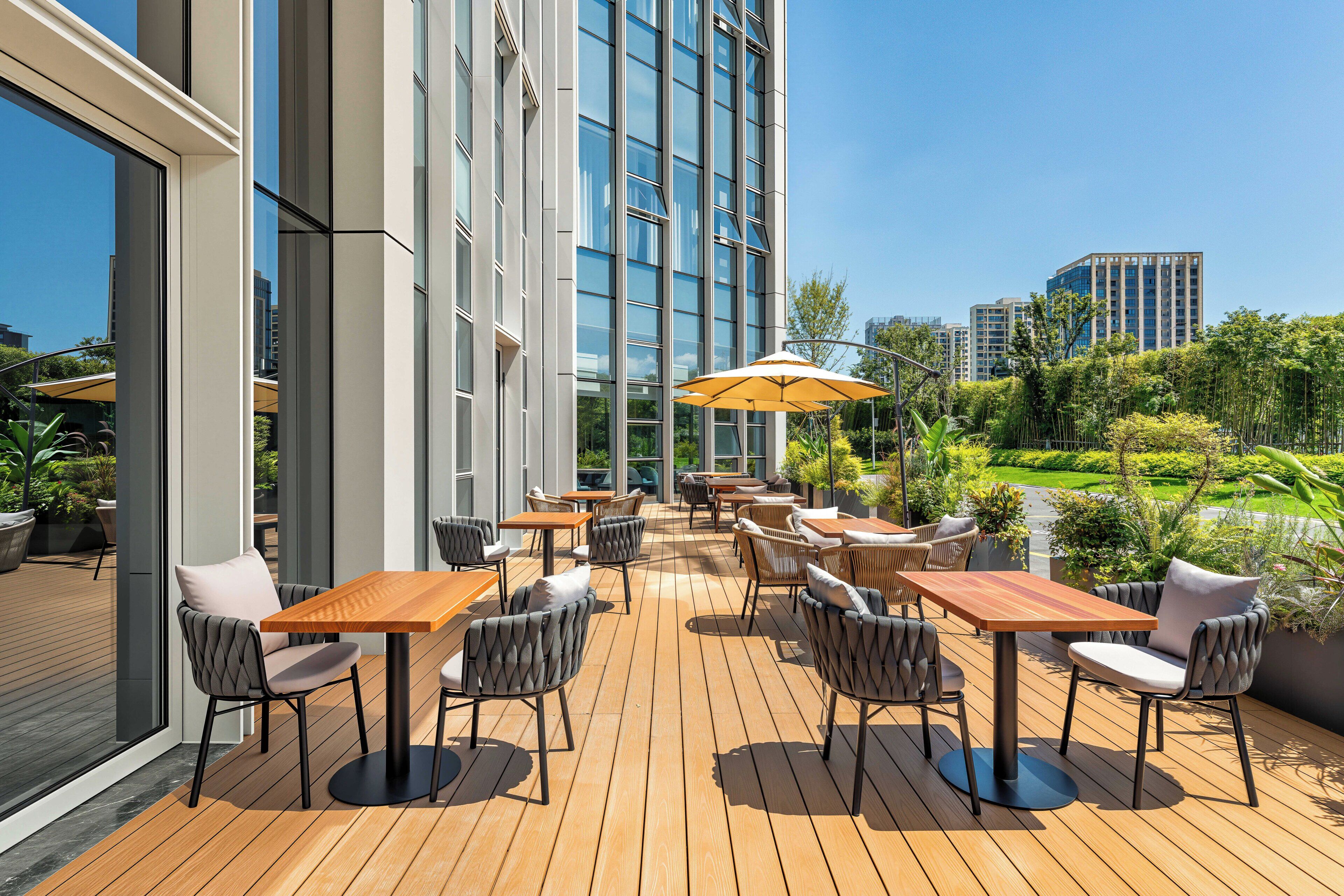 An outdoor patio with wooden tables and chairs, large windows, and a view of the cityscape.