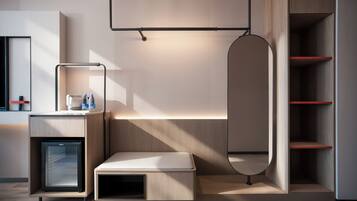 A modern hotel room with a minimalist design, featuring a built-in desk with a lamp, a small seating area, and a wardrobe with shelves.