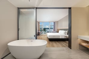 A modern bedroom with a large bathtub, a bed with multiple pillows, and a city view through large windows.