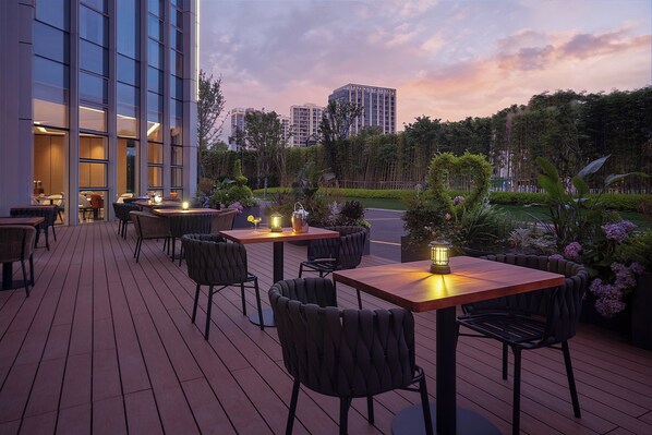 Un patio al aire libre con mesas y sillas, vista a un edificio moderno y un jardín ajardinado.