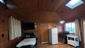 L’intérieur d’un chalet en bois comprend une table à manger, des chaises, un téléviseur, une cheminée, un réfrigérateur, un coin cuisine et une fenêtre avec des rideaux.