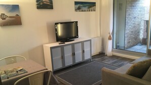 Ein Wohnzimmer mit Fernseher auf einer Kommode, einer Couch, einem Esstisch mit Stühlen und Blick durch eine Glastür.