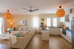 A modern living room with a sofa, coffee table, and a kitchen with a central island, white cabinetry, and a ceiling fan. - Tropical Haven — Private Resort Week, Sleeps 6 (Willemstad)
