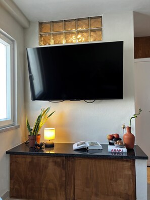 Un televisor de pantalla plana montado en una pared sobre una encimera de madera con una planta en maceta, una lámpara y un frutero.