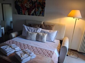 Habitación de hotel con cama, almohadas, mesita de noche con toallas, lámpara y un cuadro en la pared.