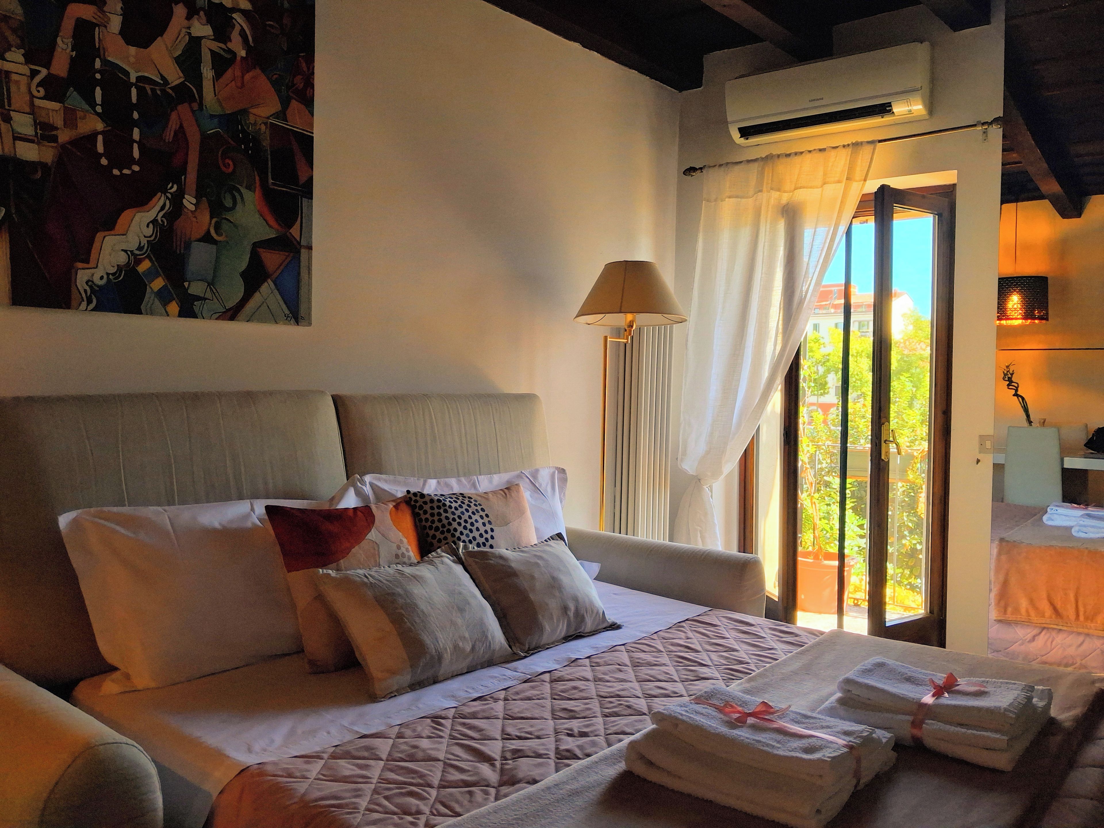 A hotel room with a bed, a window with curtains, an air conditioning unit, and a painting on the wall.
