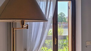 A room with a floor lamp, a window with sheer curtains, and a view of greenery outside. - Adige View Suite (Verona)
