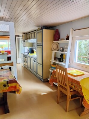 A kitchen with wooden cabinets, a dining table with chairs, and a window with a view of greenery. - Wooden mobile home on a residential leisure park access to the leisure area (La teste de buch)