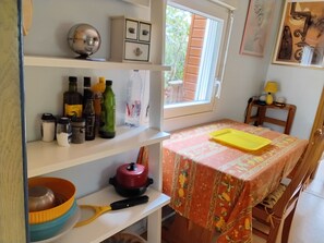 Une cuisine avec une table recouverte d’une nappe à motifs, une planche à découper jaune et divers ustensiles de cuisine rangés sur des étagères.
