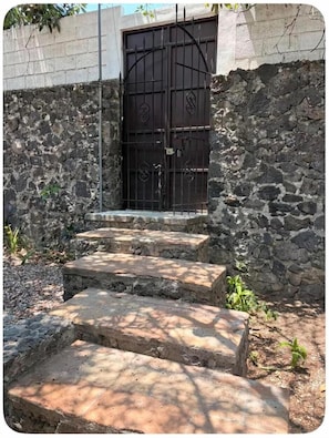 A set of stone steps leading to a metal gate with decorative patterns, surrounded by a stone wall and some vegetation. - Casa rústica en Jiutepec con alberca, jardín y asador ideal para descansar. (Jiutepec)