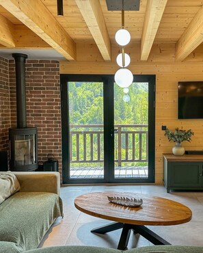 A cozy room with a fireplace, a round wooden table, and a balcony with a view of trees. - Cosy chalet with jacuzzi for 11 (Bonnevaux)