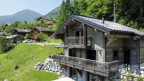 A two-story wooden house with a balcony, surrounded by greenery and mountains. - Cosy chalet with jacuzzi for 11 (Bonnevaux)