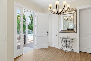 Ein Zimmer mit Holzboden, weißen Türen, einer Chandelier, einem Spiegel und einem kleinen Tisch mit Glasplatte.