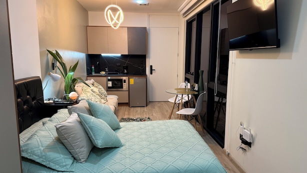 Una camera da letto moderna con un letto, cuscini, una scrivania, una sedia, un tavolino, una pianta e un angolo cottura con frigorifero e macchina del caffè.