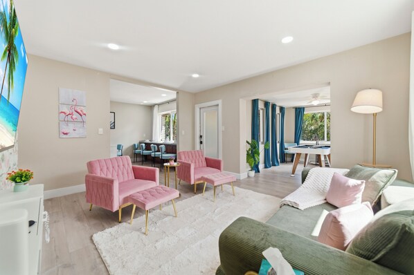 A spacious living area with a sofa, two pink armchairs, a dining area with a table and chairs, and a view of the outdoors. - Tropical Escape•Pool•Outdoor Cinema• Near Beach (Fort Lauderdale)