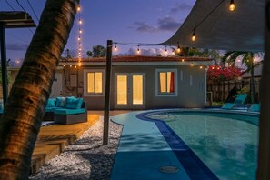 A pool area with a curved edge, a covered seating area with cushions, and string lights. - Tropical Escape•Pool•Outdoor Cinema• Near Beach (Fort Lauderdale)