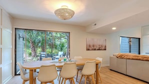 Uma área de refeições com mesa e cadeiras de madeira, luminária de teto trançada e vista para o verde através de grandes janelas.