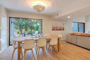 Comedor con mesa y sillas de madera, lámpara de techo tejida y vistas a follaje a través de amplias ventanas.