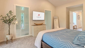 Quarto com cama, ventoinha no teto, televisão, cómoda de madeira, planta e porta de correr de vidro.