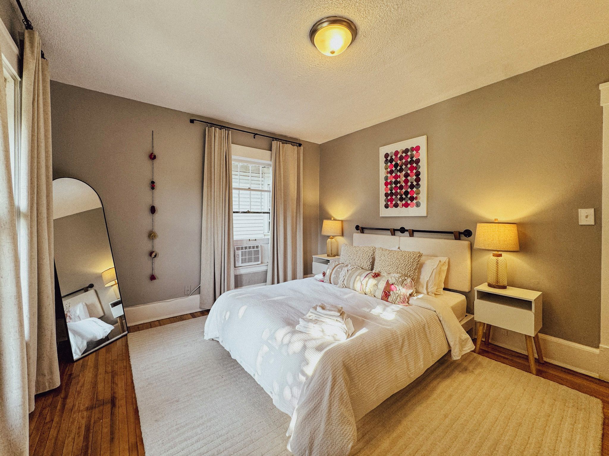 A hotel room with a large bed, bedside lamps, a mirror, a window with curtains, and a wall art piece.