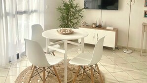 Ein modernes Wohnzimmer mit einem runden Esstisch, vier weißen Stühlen, einem großen Flachbildfernseher an der Wand, einer Bambuspflanze und einer Stehlampe.