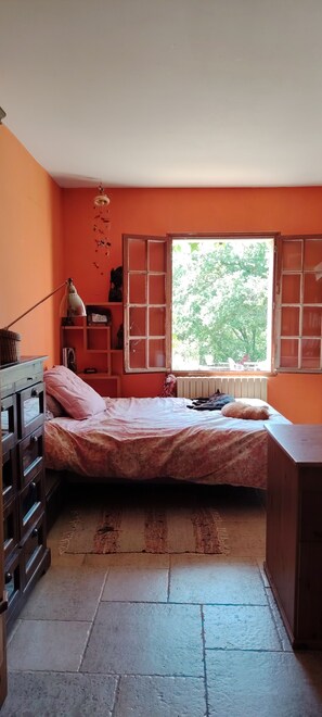 Une chambre à coucher avec un lit, une commode, une fenêtre donnant sur des arbres et un radiateur.