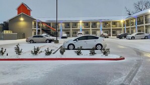 A multi-story motel with a central staircase, a parking lot with several cars, and a snow-covered ground. - Buzen Suites 2030 (Dallas)