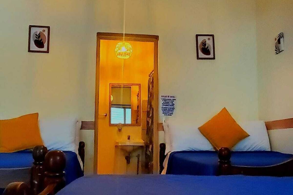 Quarto de hotel com duas camas, um candelabro, uma pia pequena e uma porta que dá para outro cômodo.