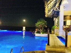 Ein Schwimmbad bei Nacht mit klarem Wasser, einer Palme und einem Gebäude mit strohgedecktem Dach.