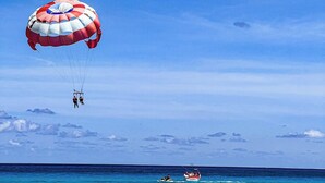 Cielo azzurro limpido con un paracadute e due persone in aria, una barca su acque turchesi calme e una costa in lontananza.