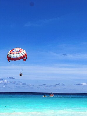 A clear blue sky with a parachute and two individuals in mid-air, a boat on calm turquoise waters, and a distant shoreline. - OCEAN FRONT APARTMENTS CANCUN (cancun)